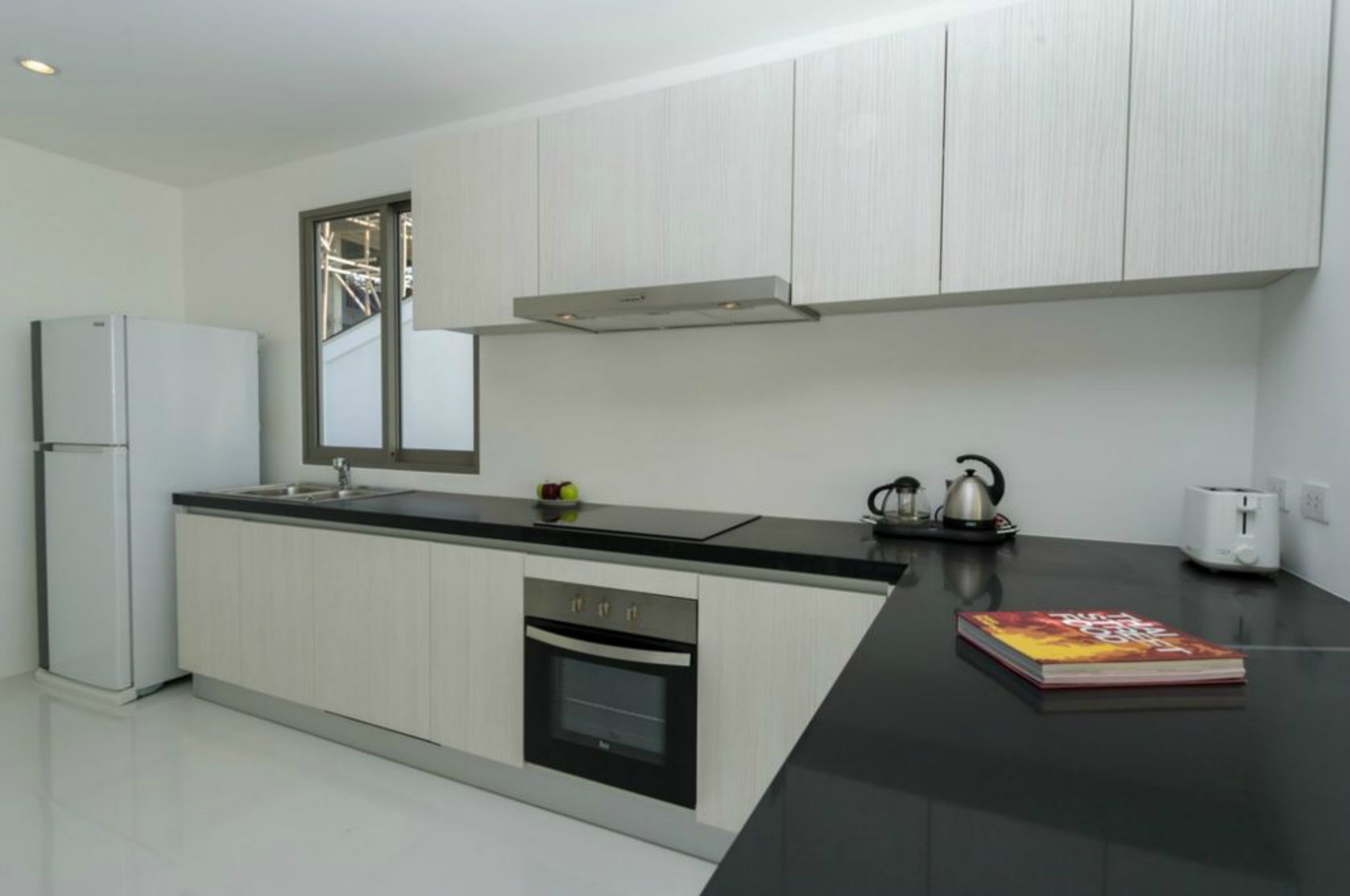 A modern kitchen with white cabinets, a black countertop, a built-in oven, a stainless steel range hood, and a kettle.