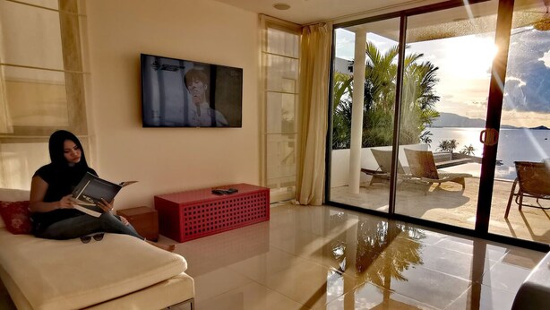 Uma sala de estar moderna com uma televisão grande, um sofá, um móvel de armazenamento vermelho e vista para uma praia através de amplas portas de vidro.