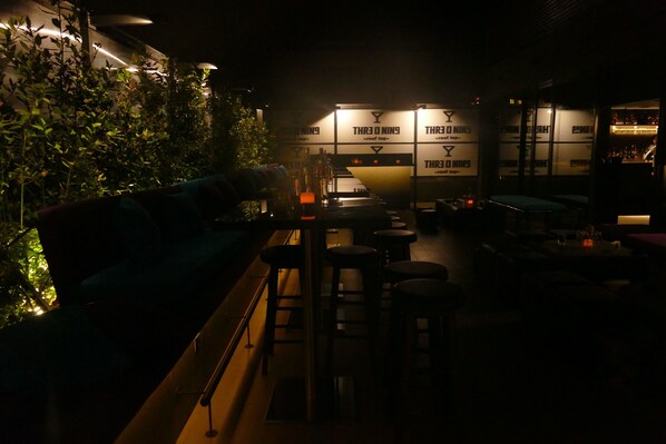 An indoor bar area with a bar counter, stools, and a seating area with a couch. There are plants on the left and a wall with illuminated signs. - Three ONine Urban House (Beirut)