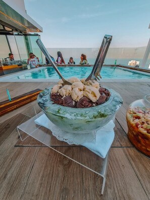 A poolside dining area with a large bowl of dessert, a glass of drink, and a view of the pool and guests. - Three ONine Urban House (Beirut)