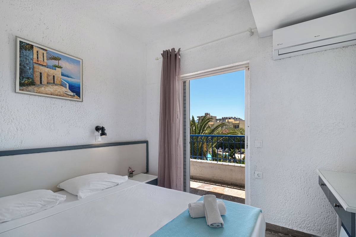 A hotel room with a bed, a painting, a window with curtains, and an air conditioning unit.