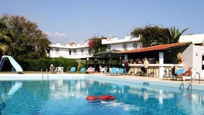 A swimming pool with lounge chairs and a bar area, surrounded by greenery and buildings. - Gorgona Hotel (Malevizi)