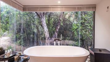 Un baño moderno con una bañera grande, una mesita con artículos de aseo y vistas a un bosque a través de amplios ventanales.
