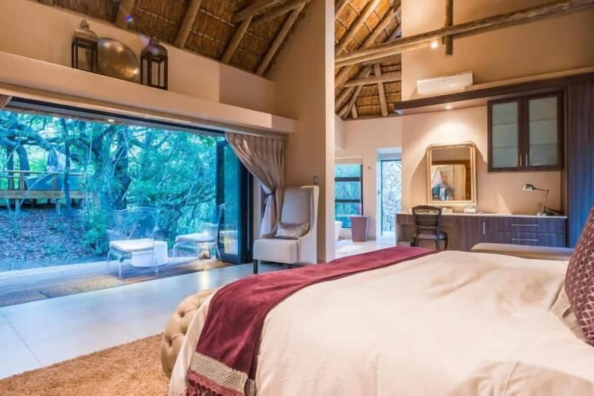 A bedroom with a large bed, a desk, and a chair. There is a window with a view of trees.