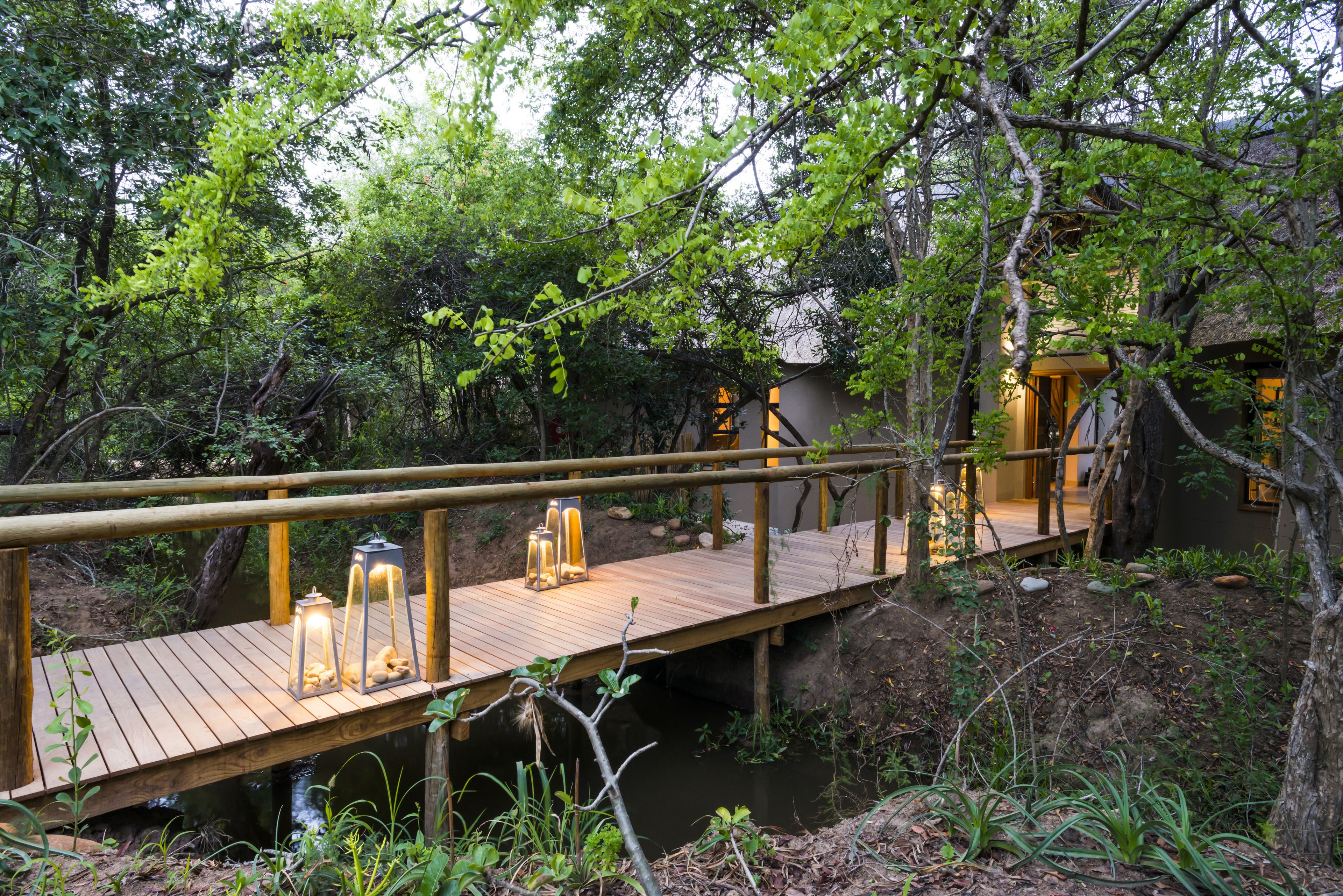 Eine hölzerne Brücke mit Laternen, umgeben von üppiger Vegetation und einem Gebäude im Hintergrund.