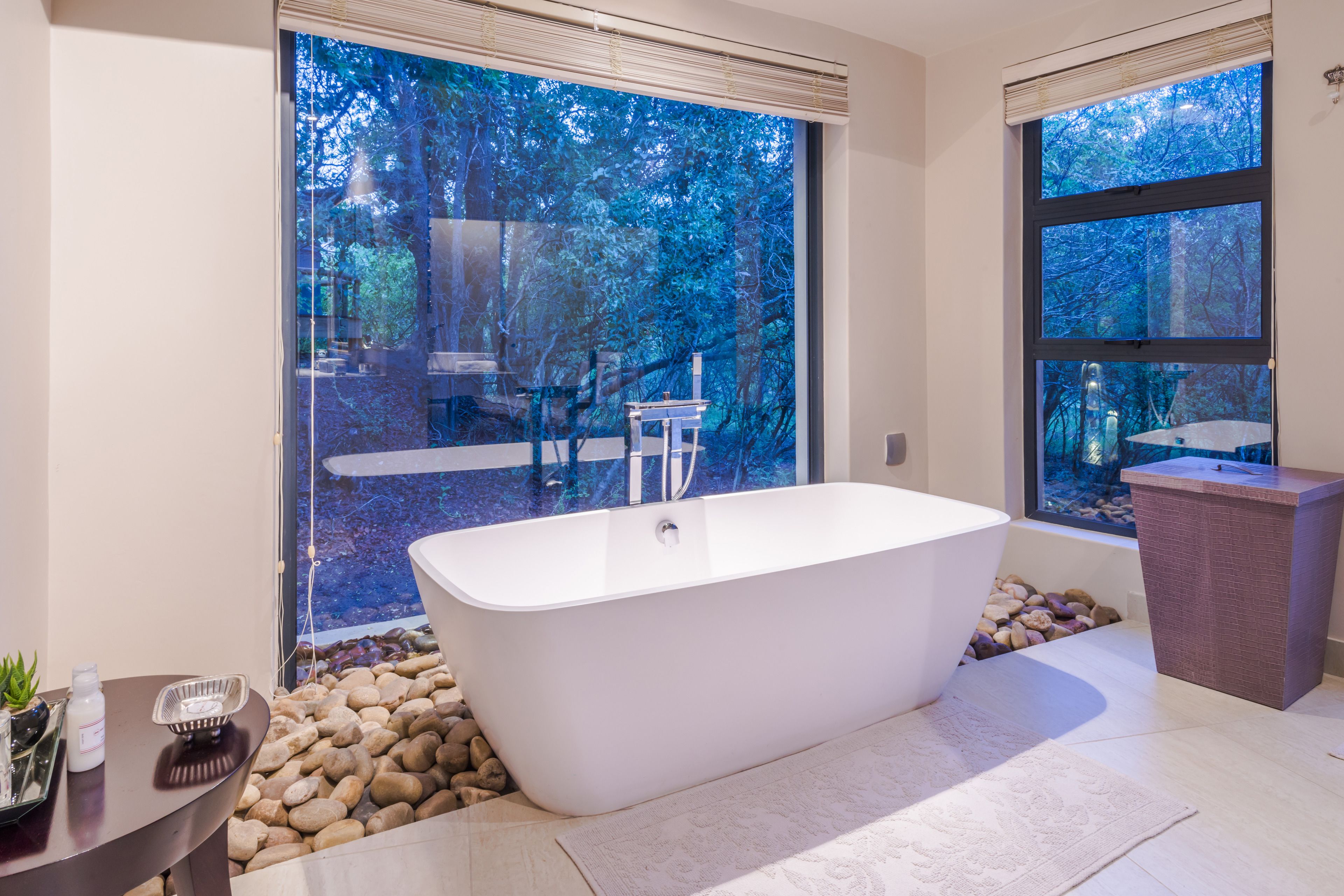 Une salle de bain moderne avec une baignoire sur pieds, de grandes fenêtres donnant sur une forêt et une petite table avec des articles de toilette.