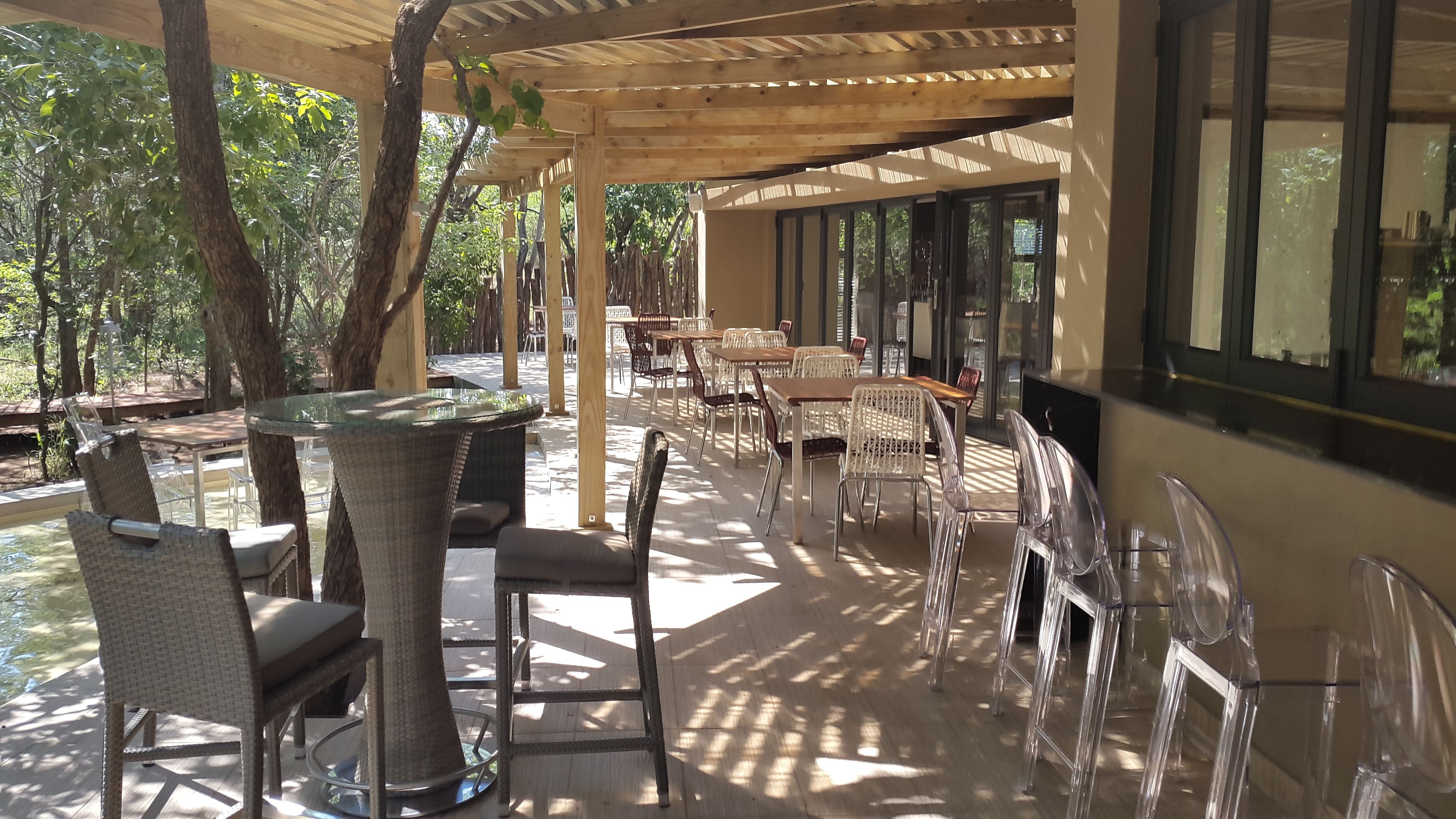 Un espace extérieur aménagé en terrasse, avec des meubles en osier et des tables en verre, entouré d’arbres et doté de grandes fenêtres.