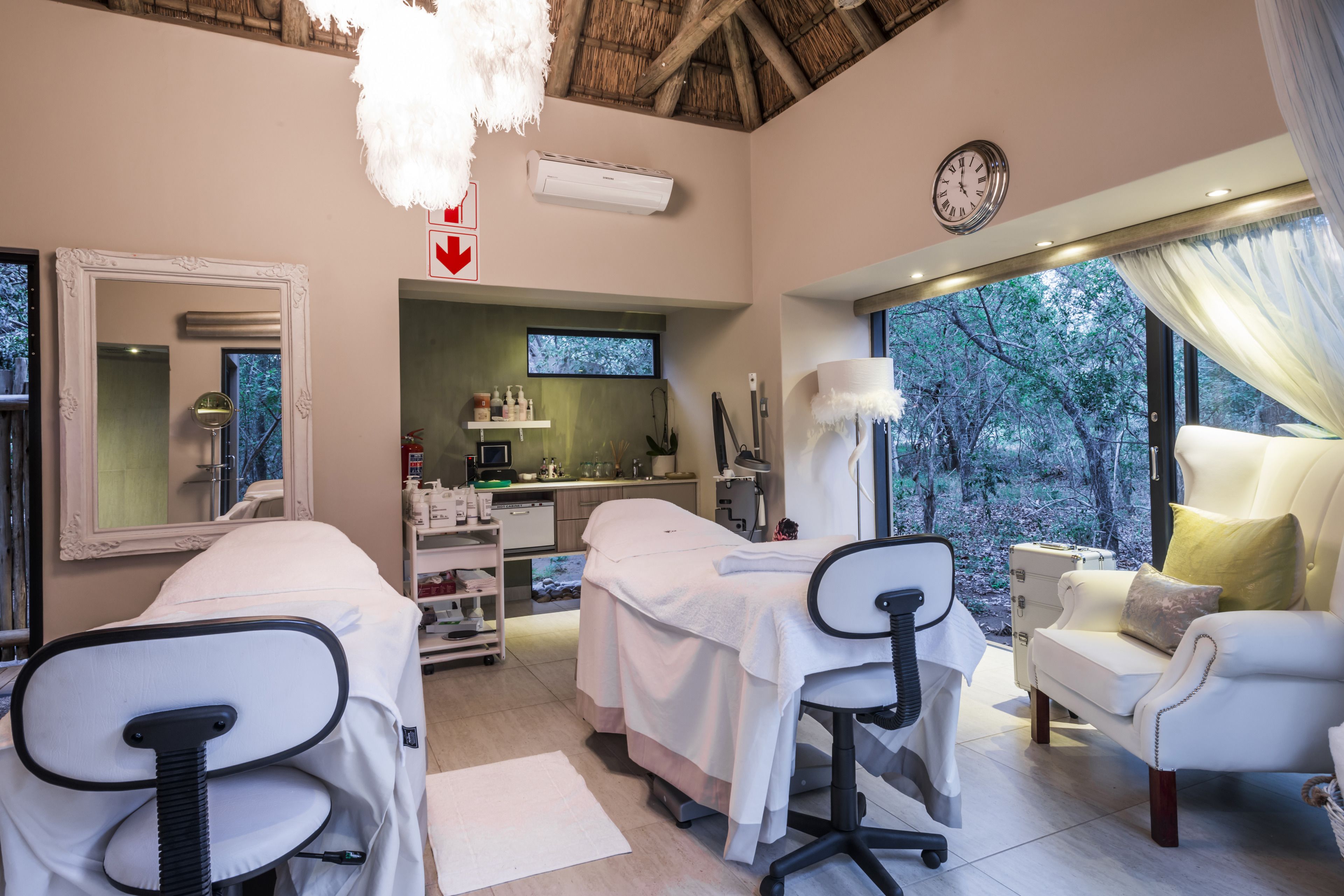 A spa room with two treatment beds, a massage table, a sofa, and a large window with a view of trees.