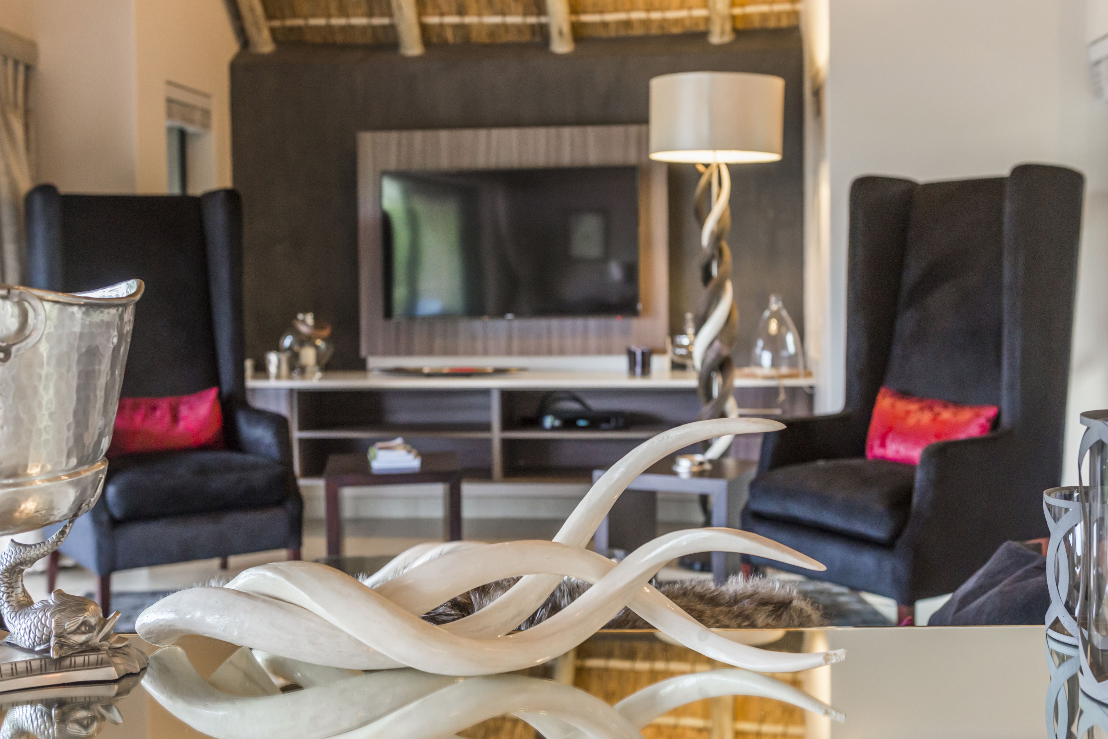 A modern living room with a large flat-screen TV, two black armchairs with red cushions, a glass coffee table, and a decorative metal sculpture.