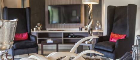A modern living room with a large flat-screen TV, two black armchairs with red cushions, a glass coffee table, and a decorative metal sculpture.