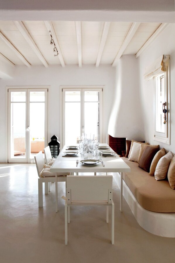 A dining area with a white table set for four, a sofa with cushions, and a view through large windows.