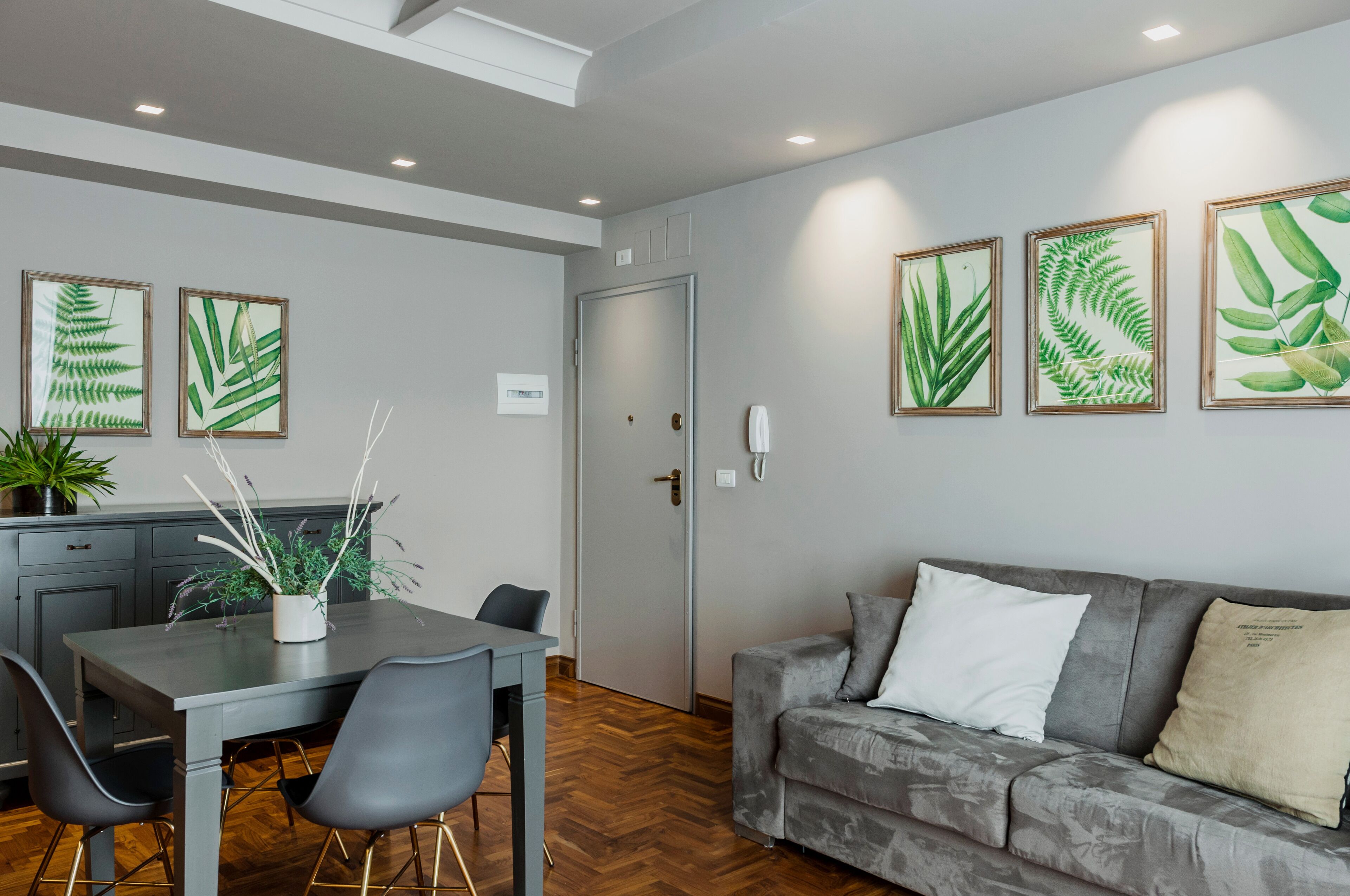 A modern living room with a grey sofa, a dining table with chairs, and framed botanical prints on the wall.