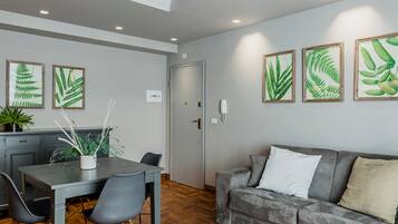 A modern living room with a grey sofa, a dining table with chairs, and framed botanical prints on the wall.