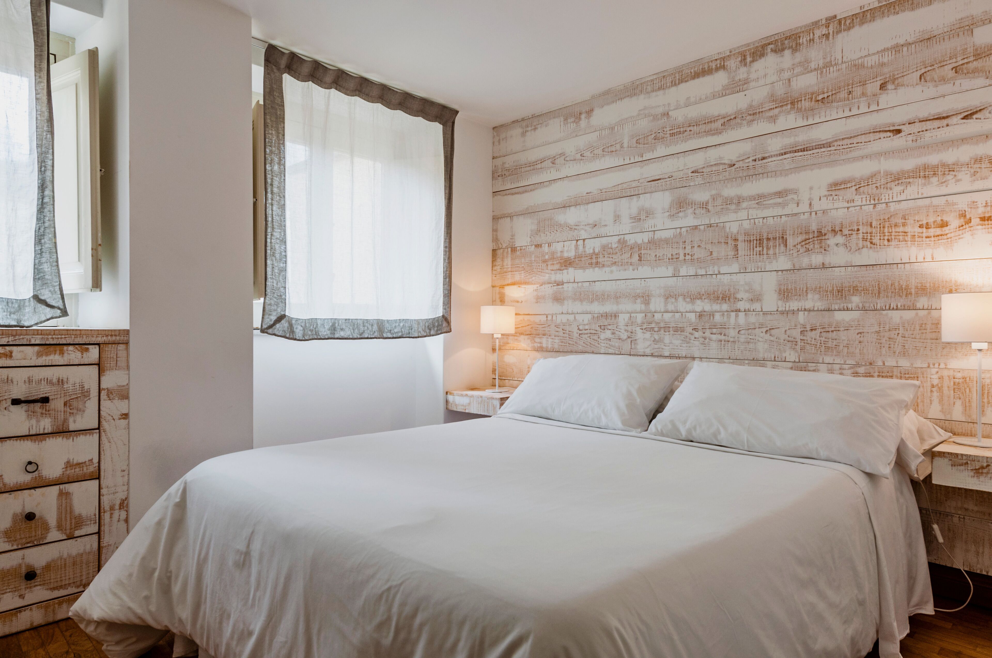 Un dormitorio con cabecera de madera, cama con ropa de cama blanca, mesita de noche con lámpara y cómoda con cajones.