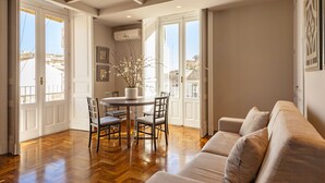 A spacious living area with a wooden floor, a round dining table with chairs, a beige sofa, and large windows offering a view of the cityscape.