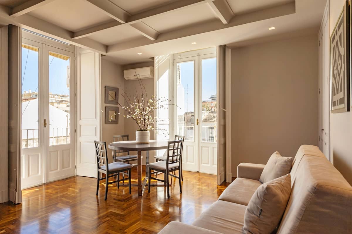 A spacious living area with a wooden floor, a round dining table with chairs, a beige sofa, and large windows offering a view of the cityscape.