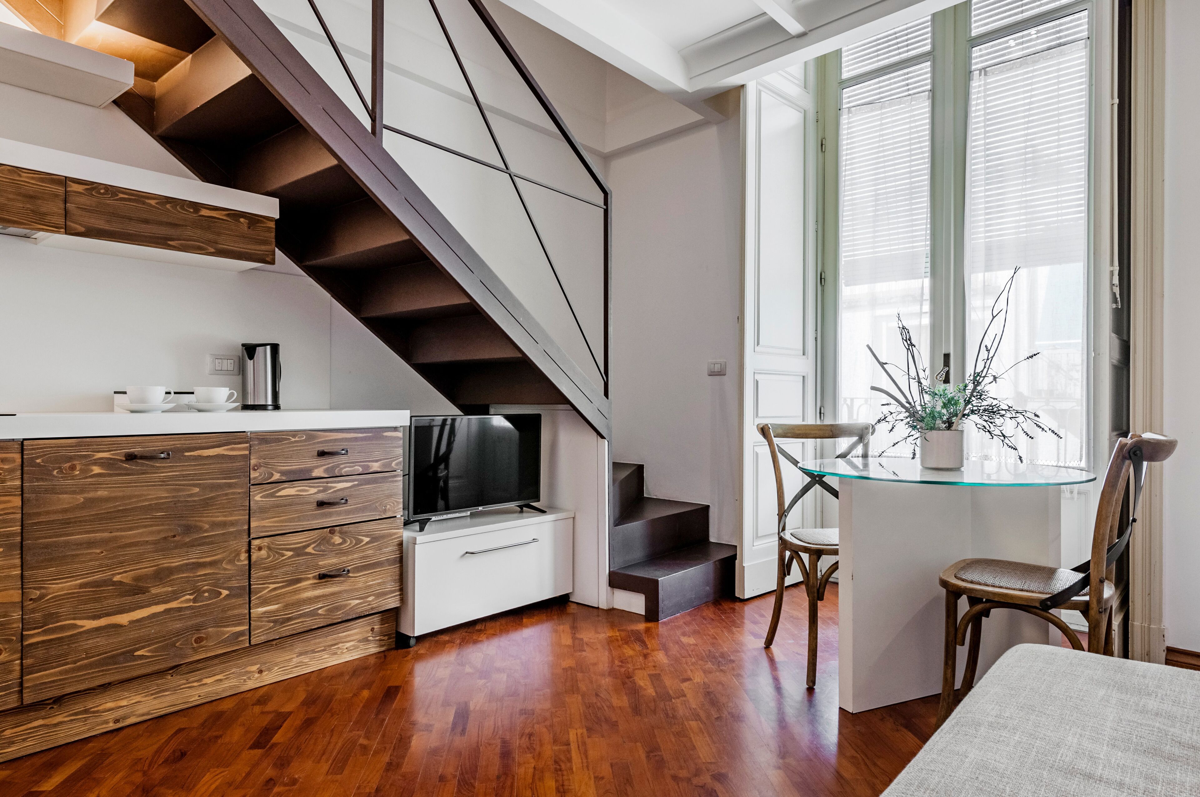 A modern kitchen with wooden cabinets, a built-in TV, and a staircase leading to a dining area with a glass table and chairs.