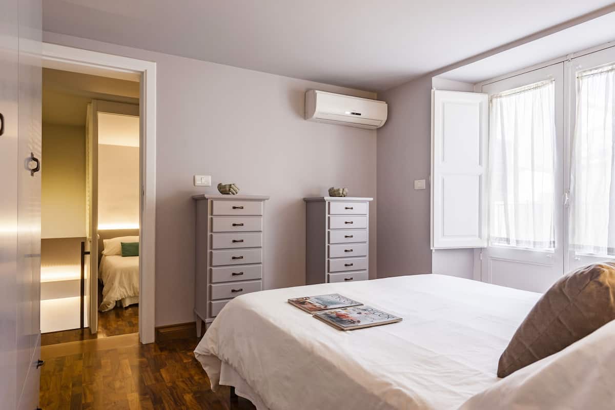 A bedroom with a bed, two dressers, an air conditioning unit, and a window with curtains.