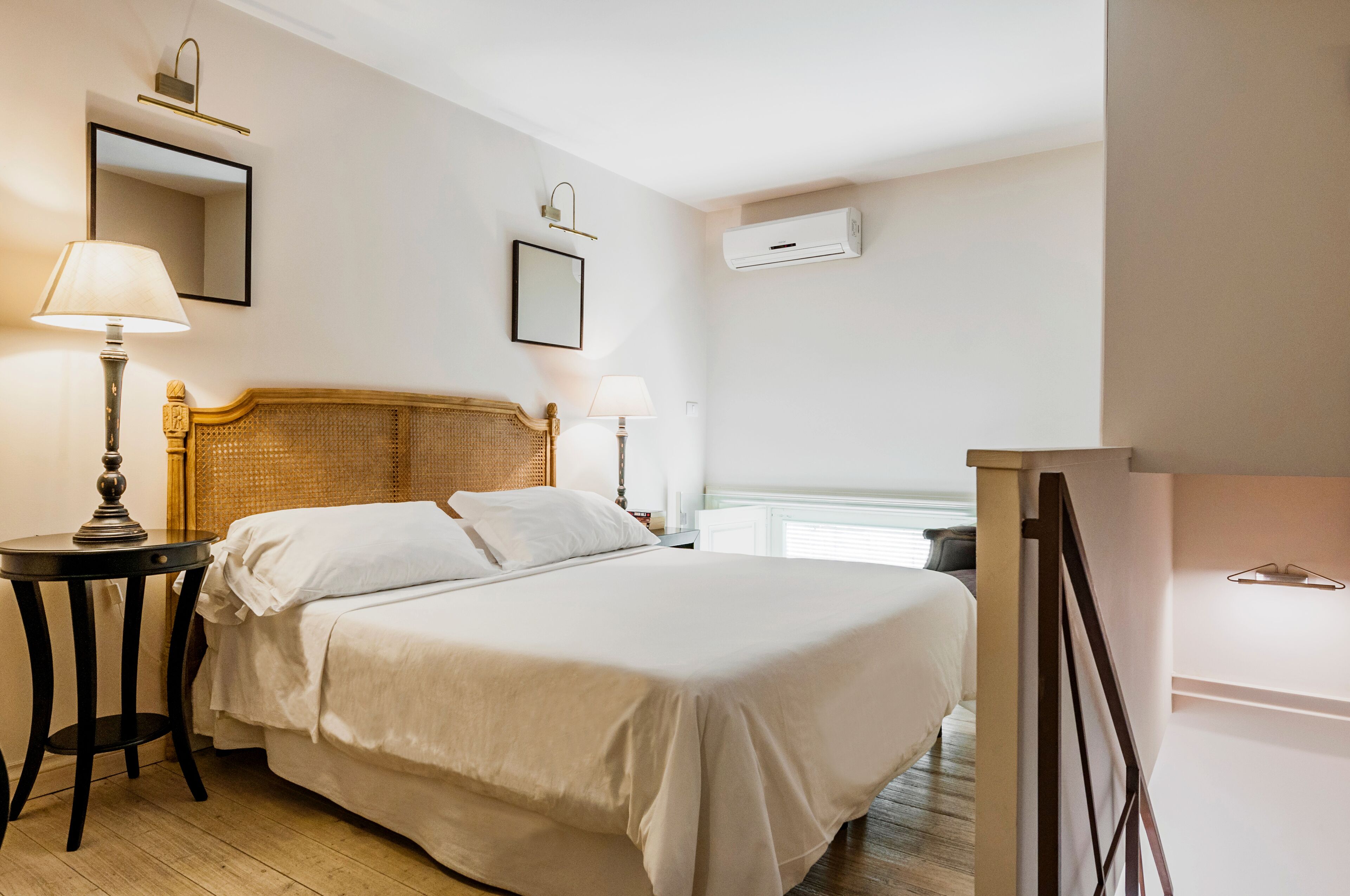 A hotel room with a bed, bedside tables, a lamp, a mirror, and an air conditioning unit.