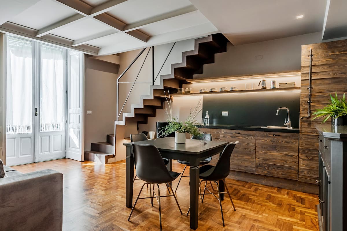 A modern kitchen with a dark countertop, wooden cabinets, and a built-in stove. There is a dining area with a table and chairs, and a staircase leading to an upper level.