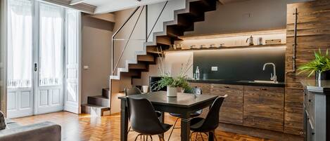 A modern kitchen with a dark countertop, wooden cabinets, and a built-in stove. There is a dining area with a table and chairs, and a staircase leading to an upper level.
