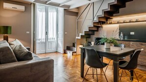 A modern living room with a sofa, a dining table with chairs, a staircase, and a kitchen area.