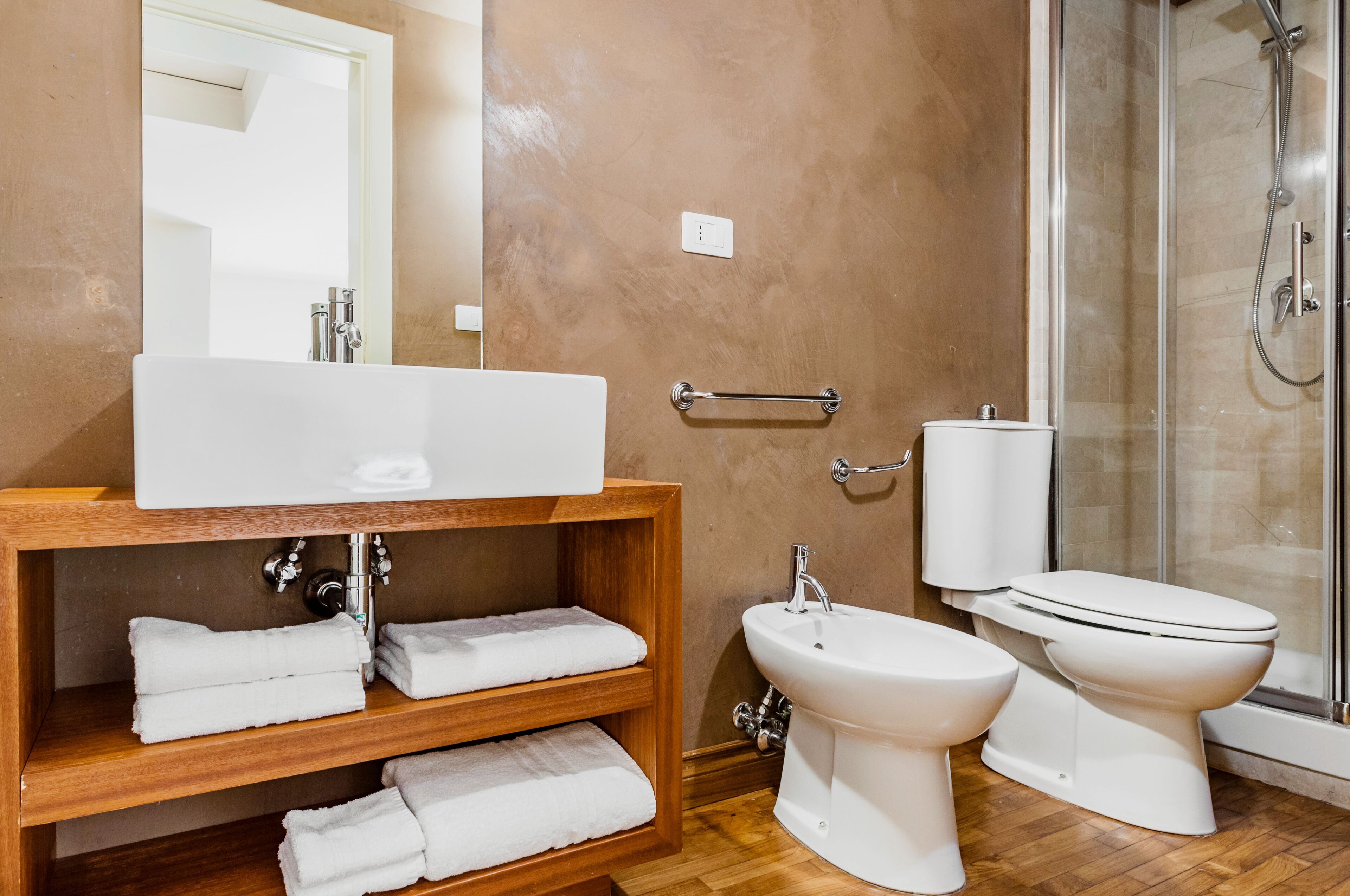 Un baño con tocador de madera, un espejo grande, un lavabo blanco, un bidé, un inodoro y una cabina de ducha con mampara de vidrio.