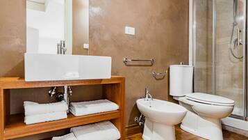 Un baño con tocador de madera, un espejo grande, un lavabo blanco, un bidé, un inodoro y una cabina de ducha con mampara de vidrio.