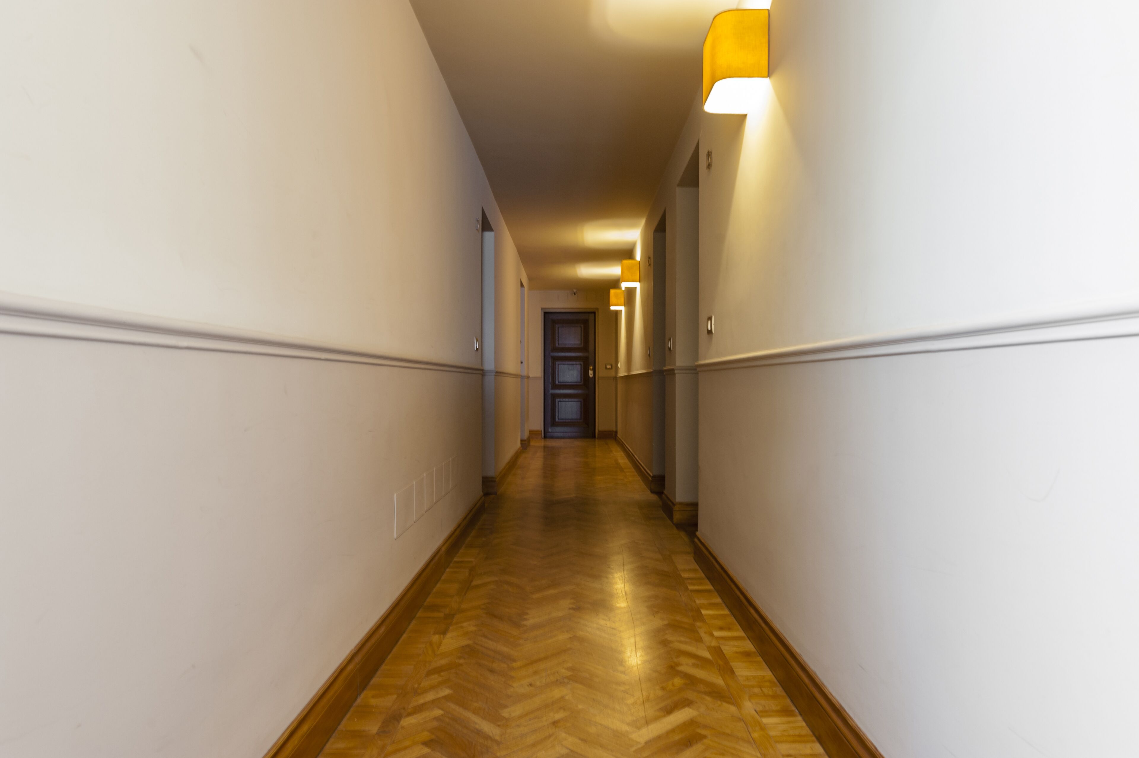 Un long couloir étroit, doté d’un plancher en bois et de murs blancs, avec une porte au bout et des luminaires fixés au mur.