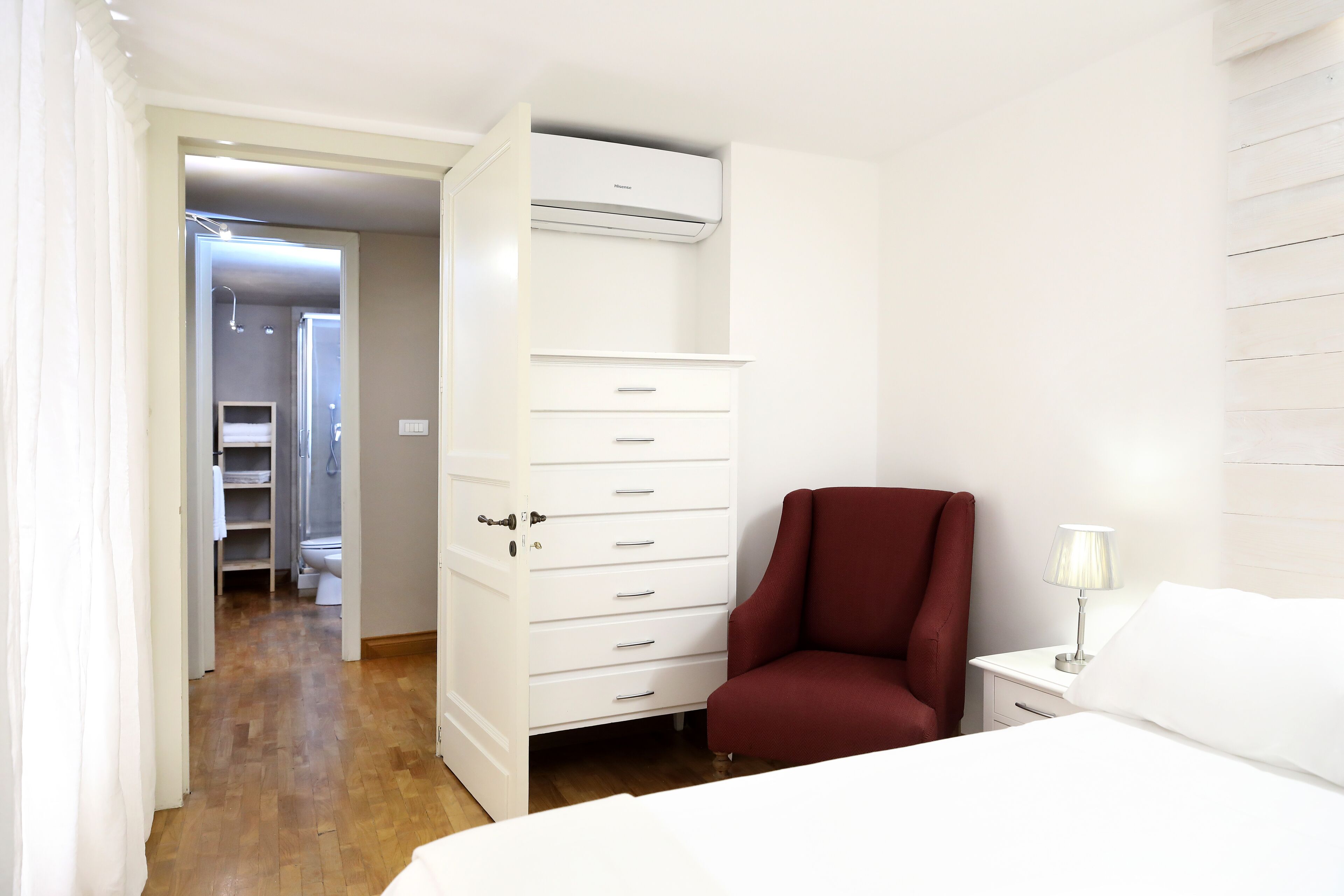 A hotel room with a wardrobe, a red chair, a bedside table with a lamp, and a bed.