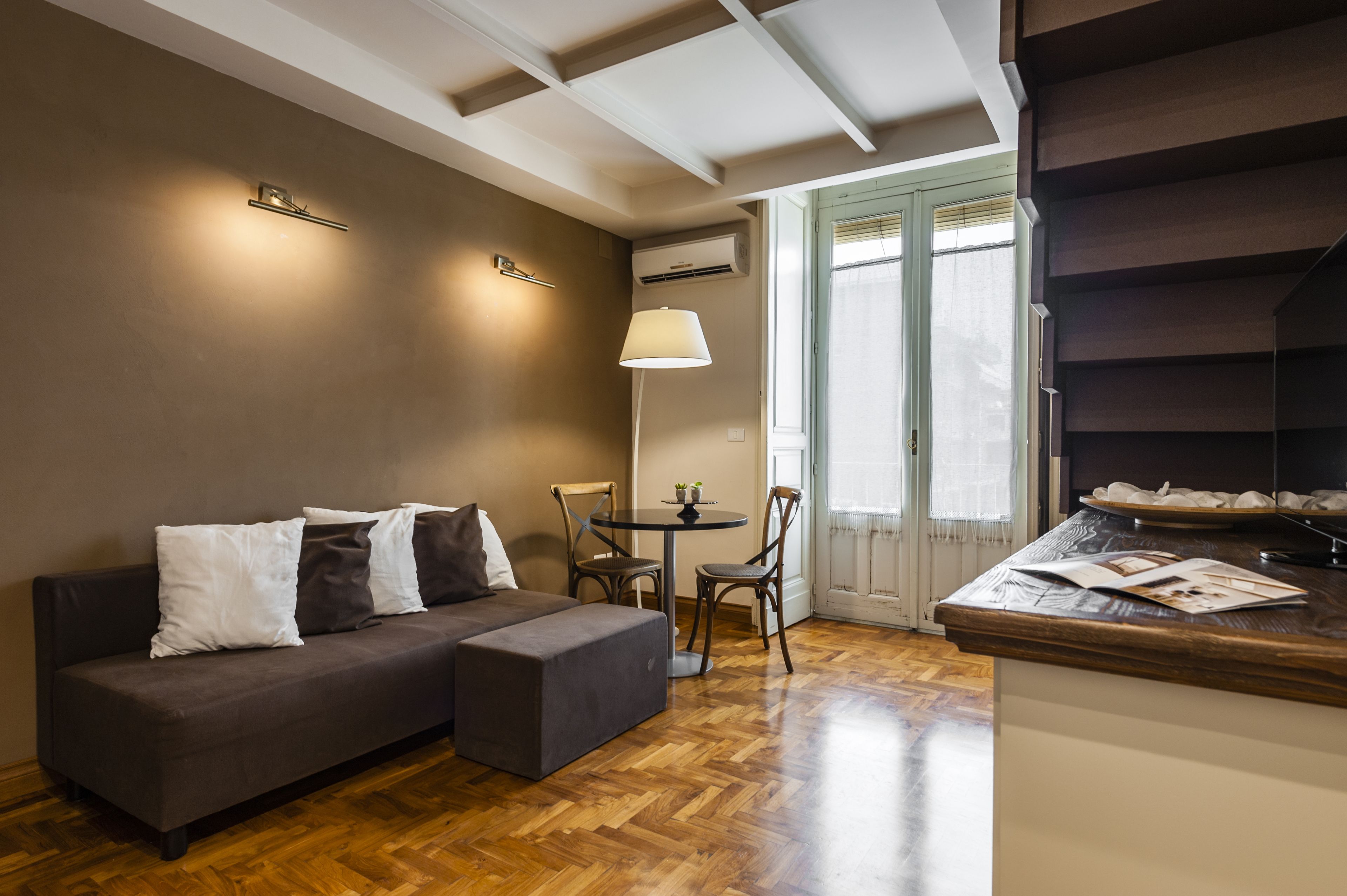 Un salon moderne avec un canapé, une petite table et des chaises. On y trouve une lampe, une fenêtre avec des stores et un plancher en bois.