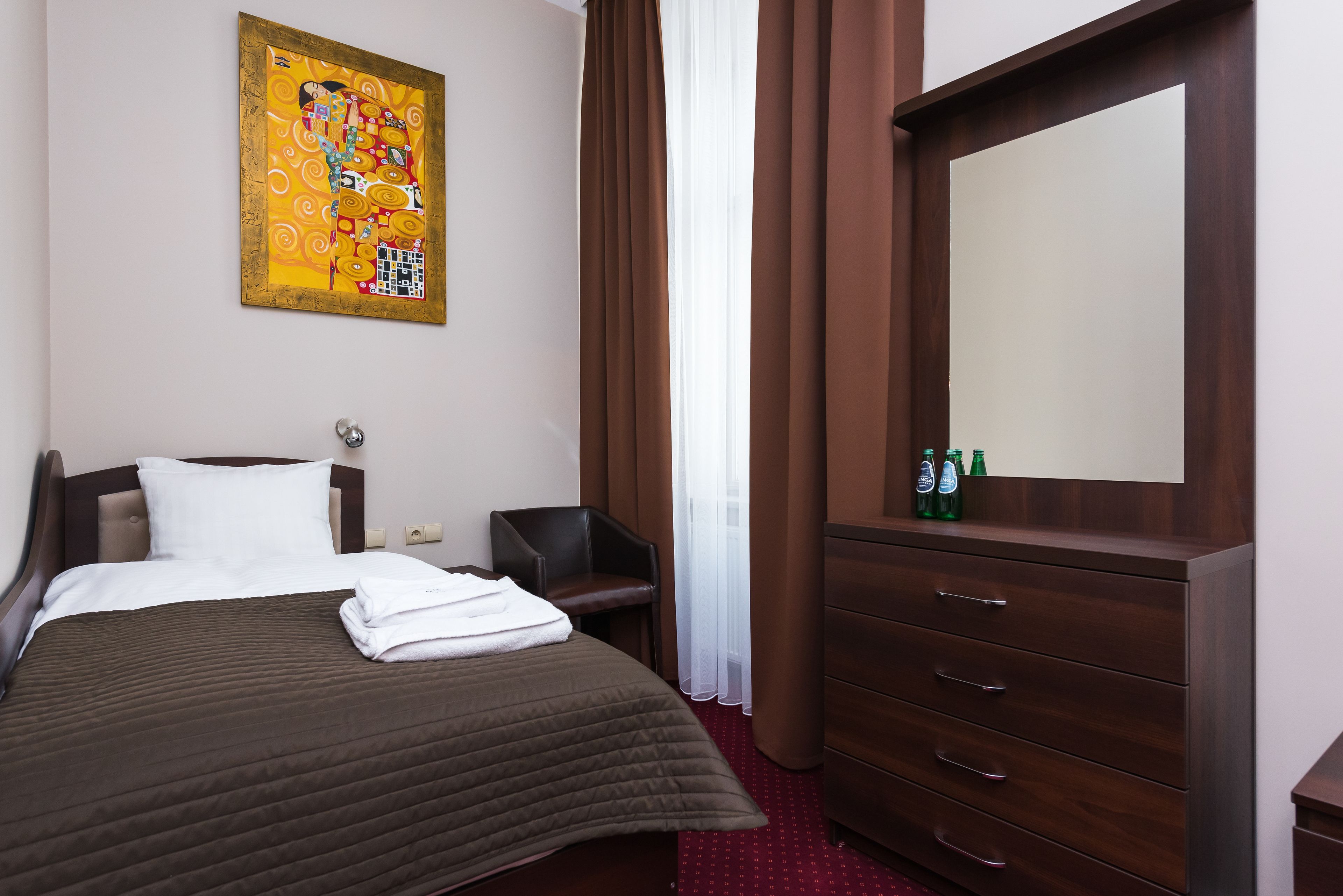 Une chambre d’hôtel avec un lit, une commode, une chaise, un tableau au mur et une fenêtre avec des rideaux.