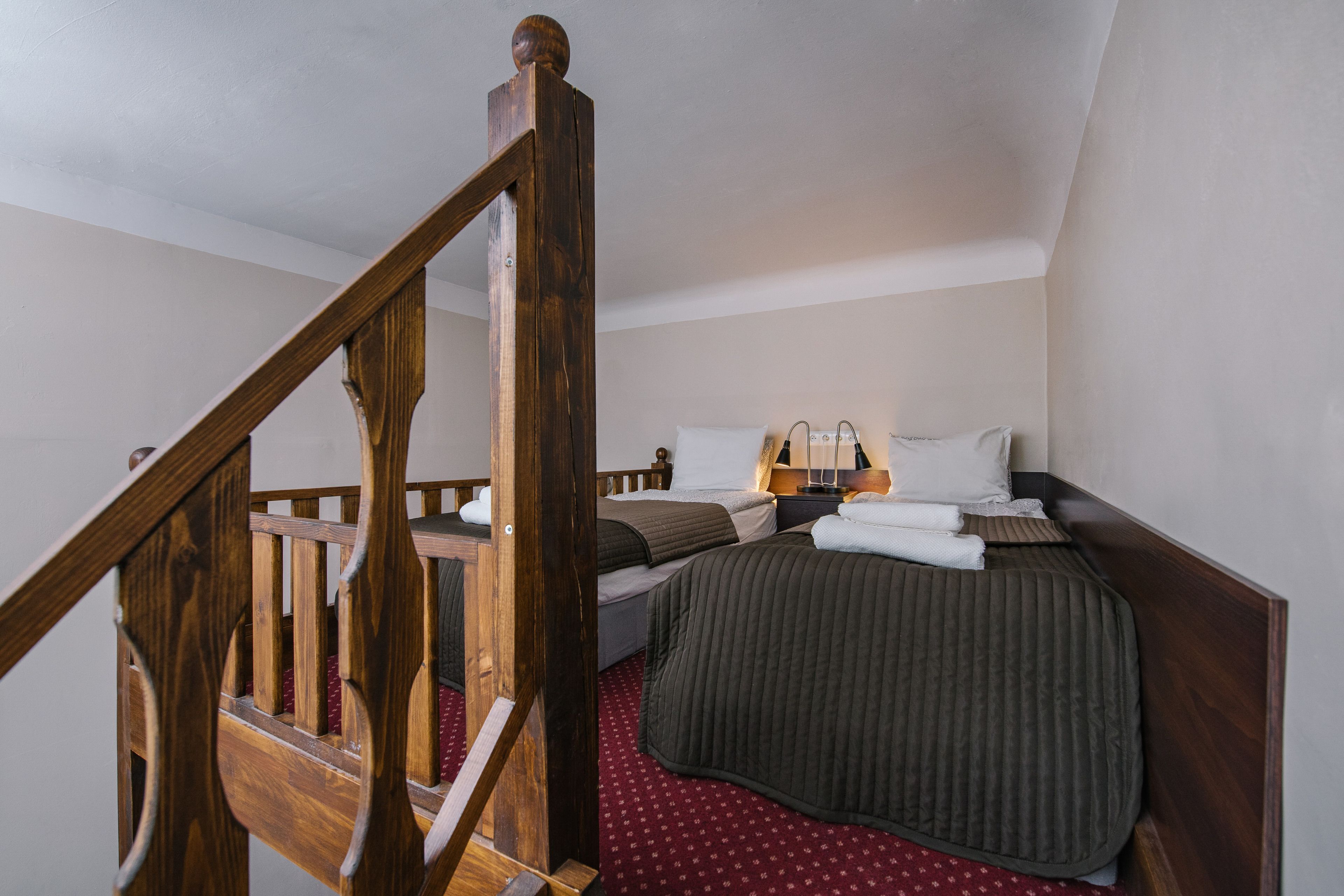 A bedroom with a wooden staircase, a bed with a dark cover, a bedside table with a lamp, and a red patterned carpet.
