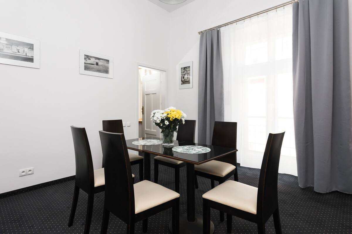 A hotel room with a dining table, four chairs, a vase with flowers, and a window with curtains.