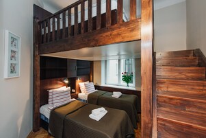 A room with a wooden bunk bed, two single beds, a window with a view, and a staircase.