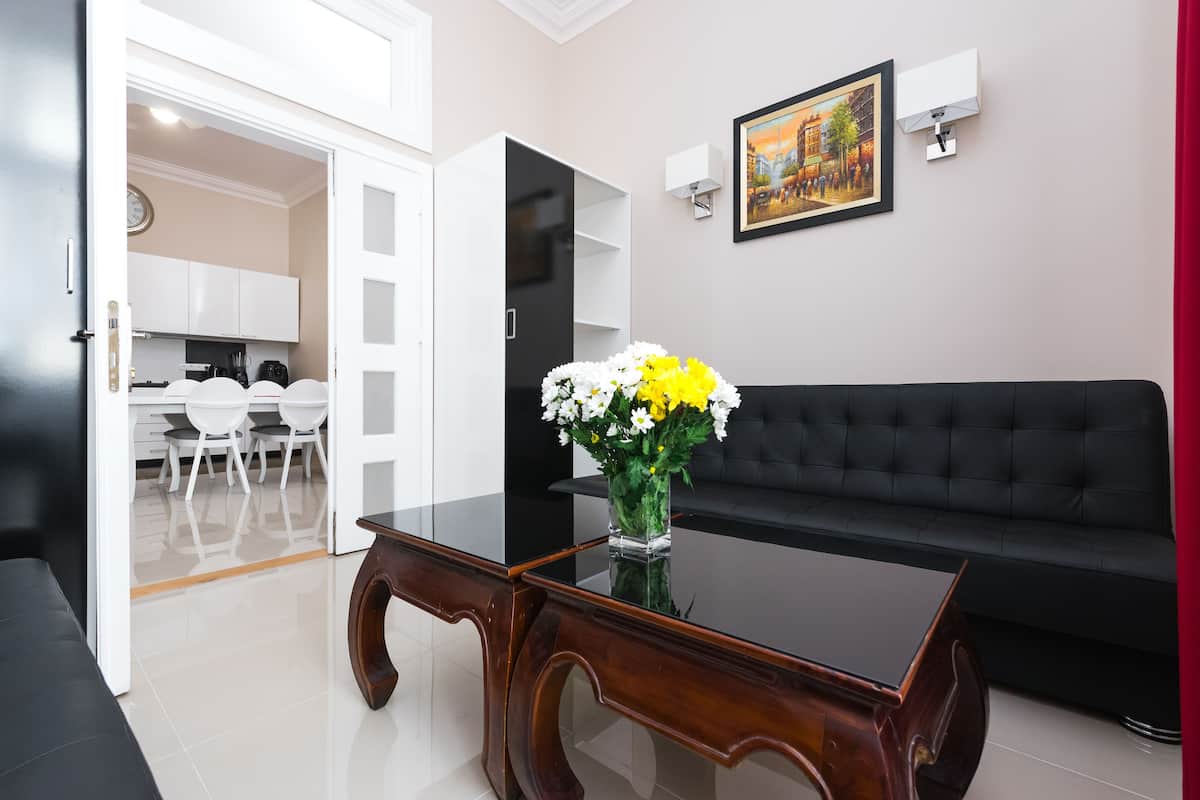 A modern living room with a black sofa, a wooden coffee table with a vase of flowers, a dining area with white chairs, and a kitchen in the background.