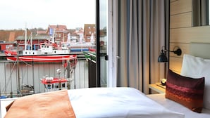 A hotel room with a bed, a nightstand, a lamp, and a view of boats and buildings through a window. - Hafenhotel Meereszeiten (Heiligenhafen)