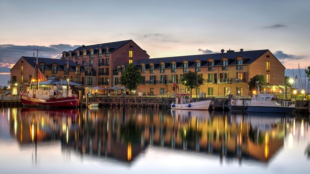 A waterfront hotel with multiple balconies, a dock with boats, and a calm water reflection. - Hafenhotel Meereszeiten (Heiligenhafen)