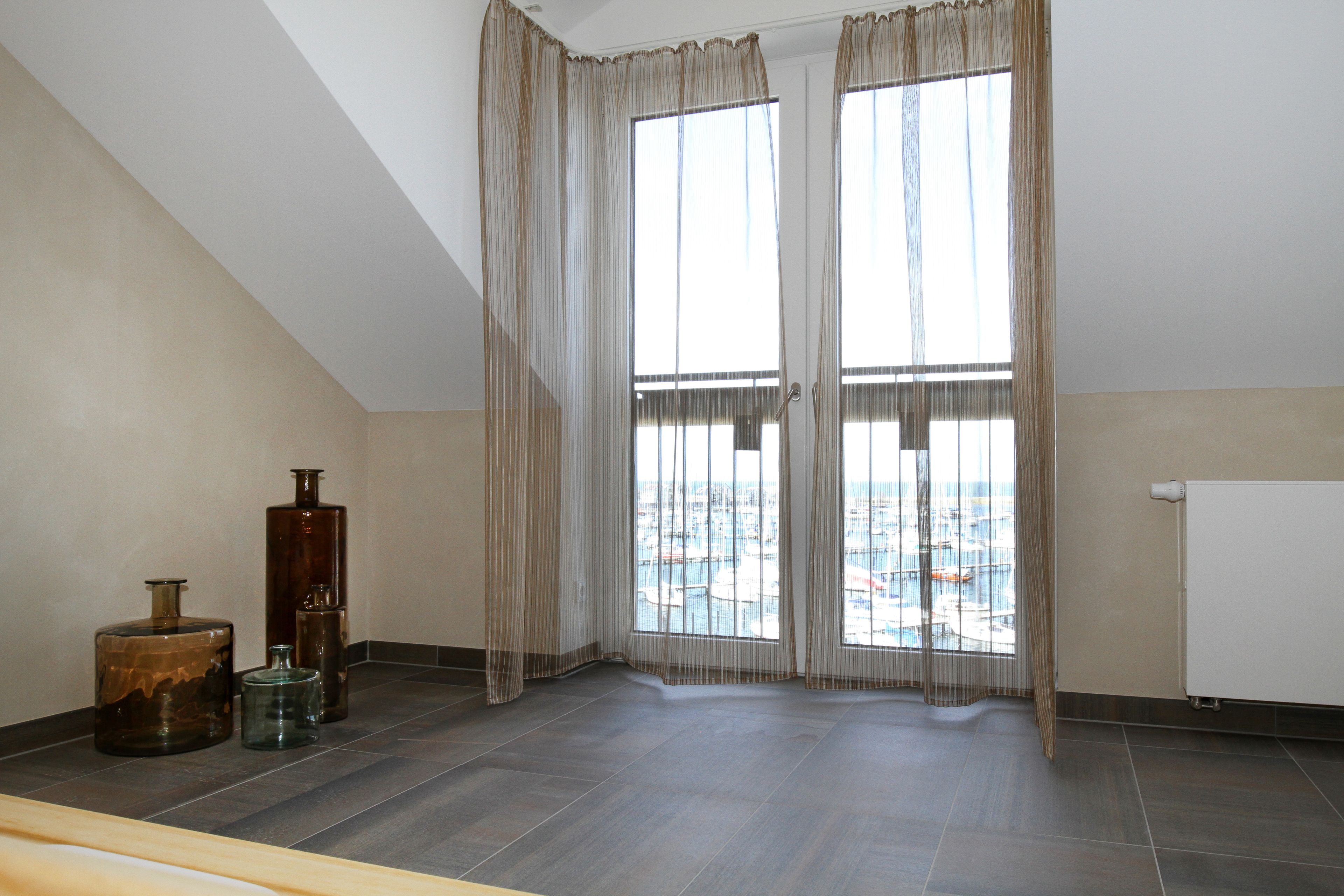 A room with a sloped ceiling, large windows with sheer curtains, and a view of a cityscape. There are three large vases on the floor.