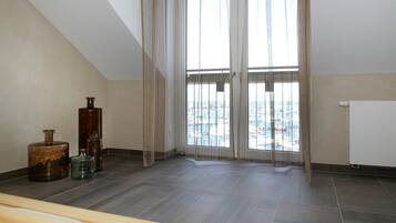 A room with a sloped ceiling, large windows with sheer curtains, and a view of a cityscape. There are three large vases on the floor.