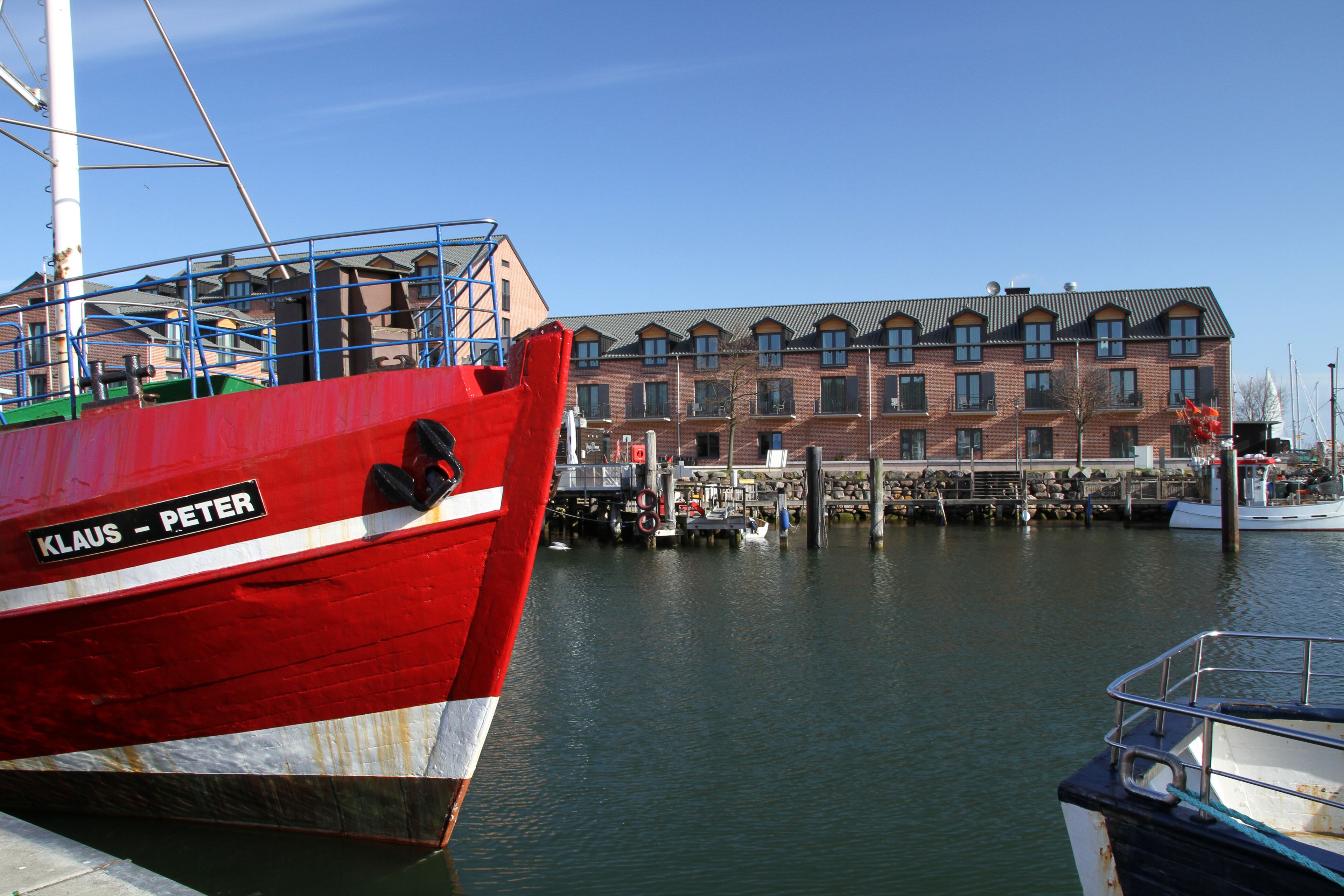 Un barco rojo llamado "Klaus - Peter" atracado en un puerto deportivo, con un edificio de ladrillo al fondo.