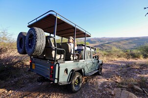 Veículo de safari com suporte no teto e pneus sobressalentes, estacionado em terreno rochoso com paisagem montanhosa ao fundo.