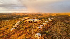 A cluster of houses with a central building, surrounded by smaller structures, set in a hilly, arid landscape. - River Crossing Lodge (Windhoek)