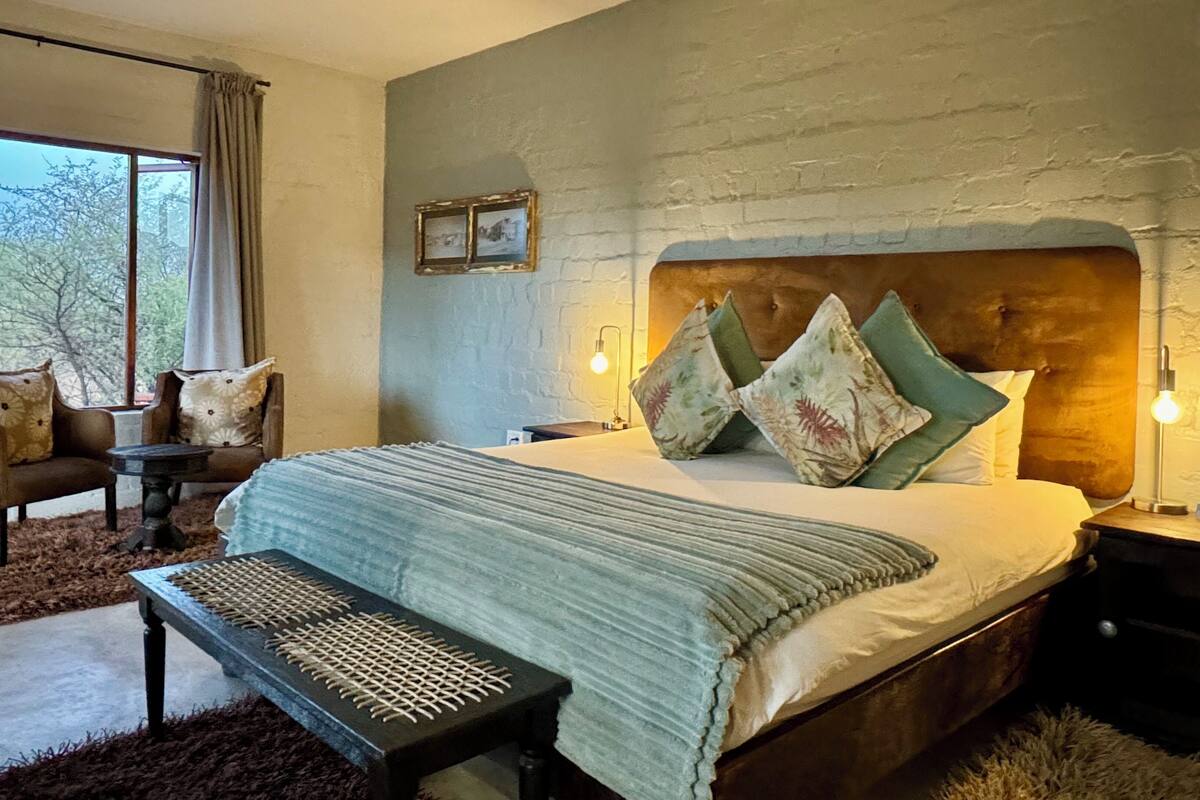 A bedroom with a large bed, a bench, a chair, and a nightstand with a lamp.