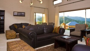 A spacious living room with a leather sofa, matching ottoman, and a coffee table. There is a bookshelf, a chandelier, and large windows offering a mountain view. - River Crossing Lodge (Windhoek)