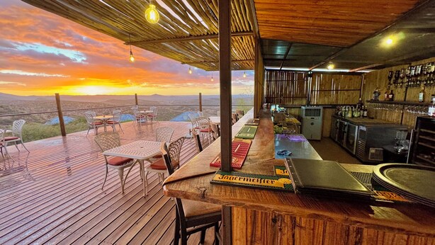 A bar area with a view of the sunset, wooden deck seating, and a bar counter. - River Crossing Lodge (Windhoek)