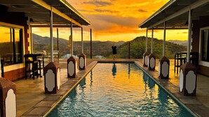 A pool with a view of mountains at sunset, surrounded by outdoor seating and a covered area. - River Crossing Lodge (Windhoek)