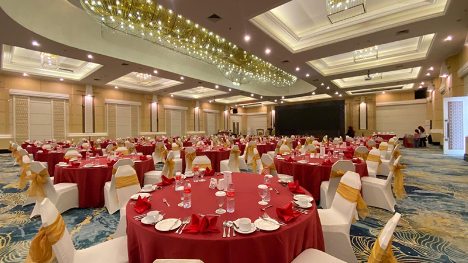 Een feestzaal met ronde tafels gedekt voor een formeel evenement, met witte stoelen met gouden linten, rode tafelkleden en een podium aan de voorzijde.