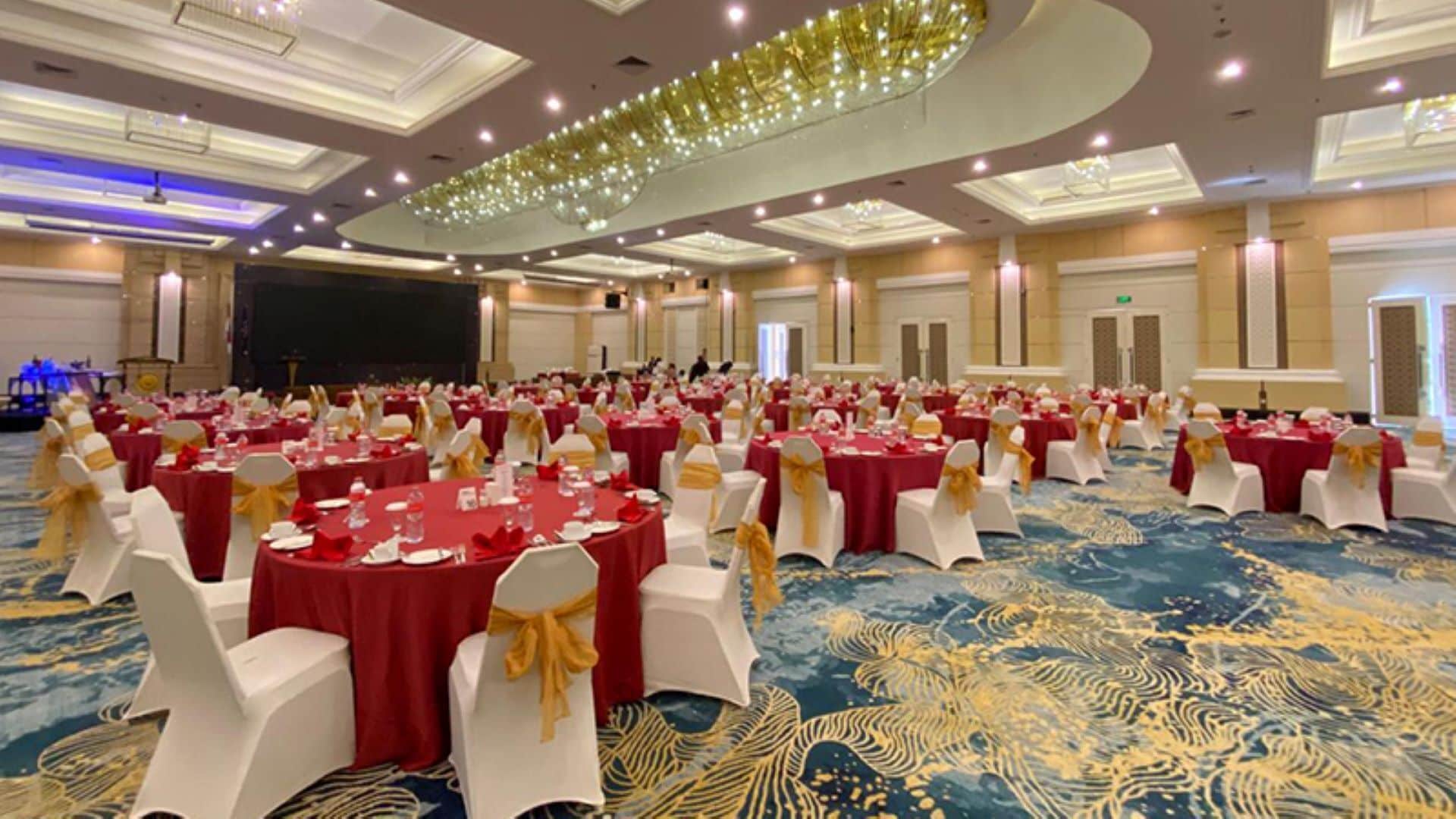 Une vaste salle de banquet avec des tables rondes recouvertes de nappes rouges et des chaises blanches ornées de rubans dorés.