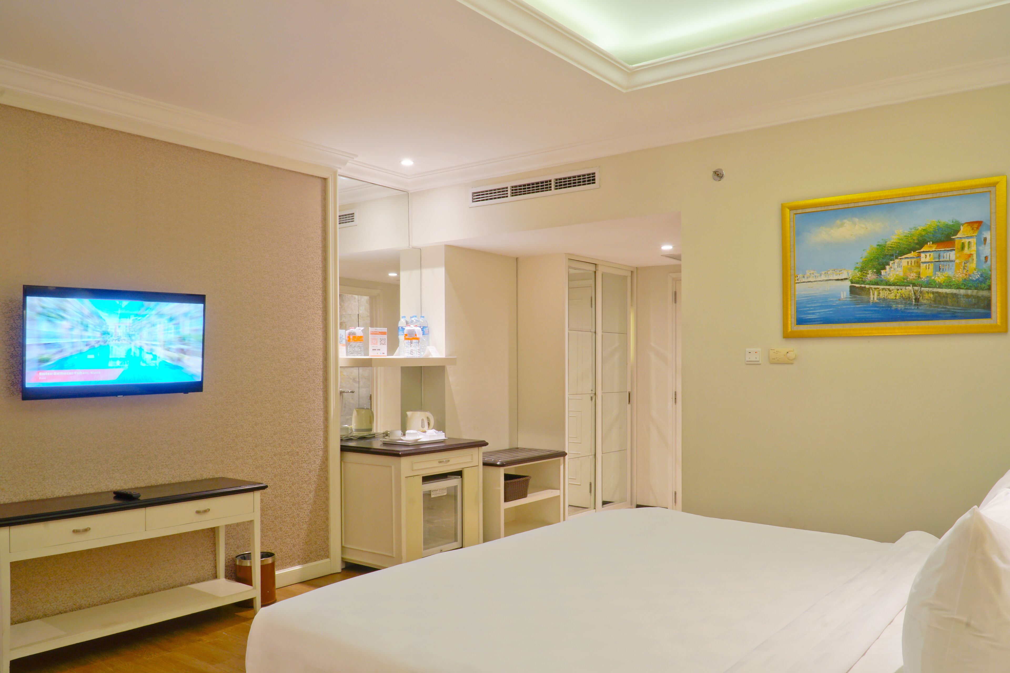 A hotel room with a bed, a TV mounted on the wall, a desk with a computer, a small kitchenette, and a painting on the wall.